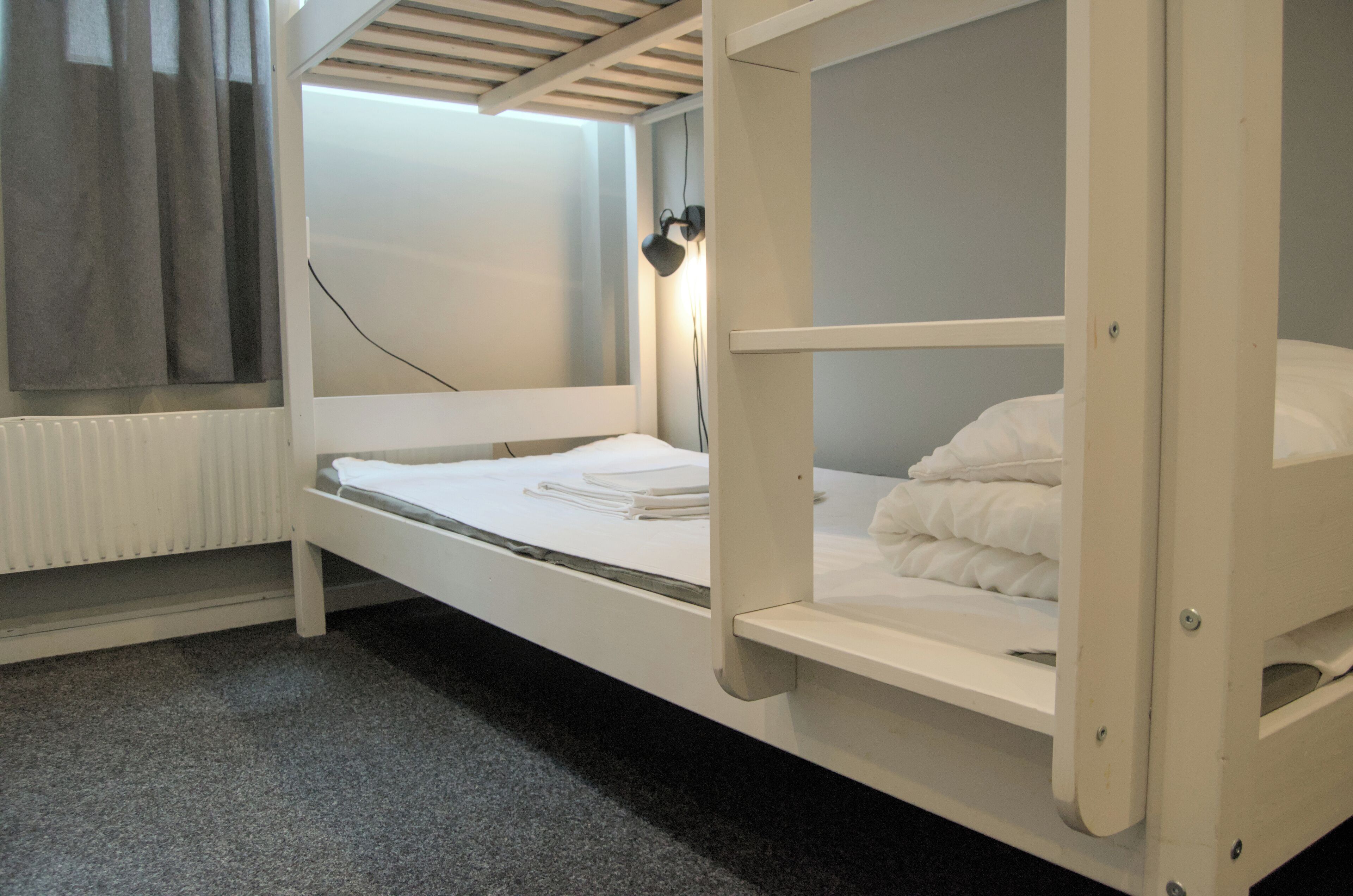 Una cama litera con estructura blanca, una mesita de noche, una lámpara y una ventana con cortinas.