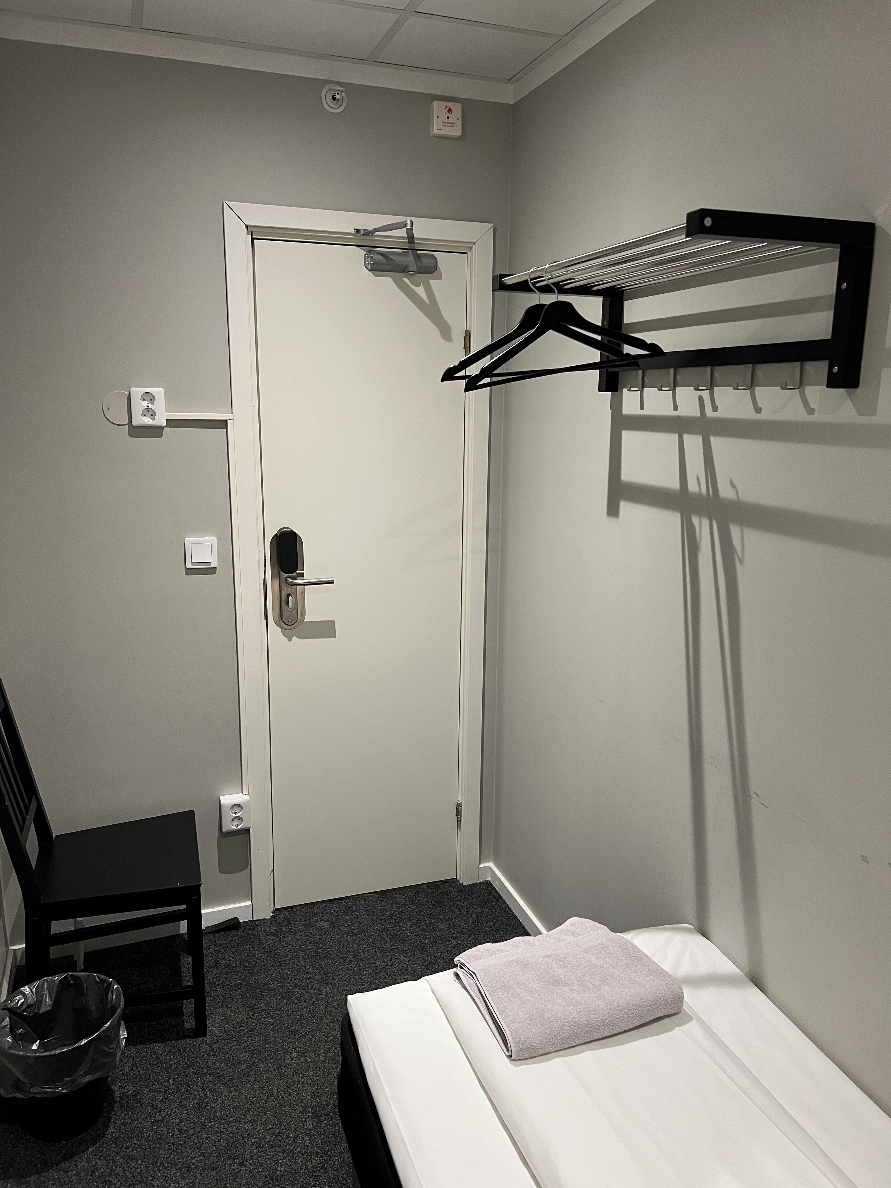 A hotel room with a bed, a chair, a coat rack, and a door.