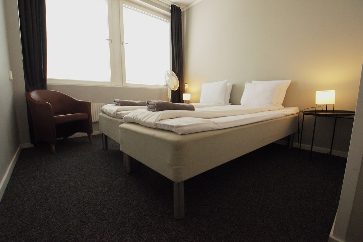 Ett hotellrum med en säng, en stol, ett sängbord, en lampa och ett fönster med gardiner.