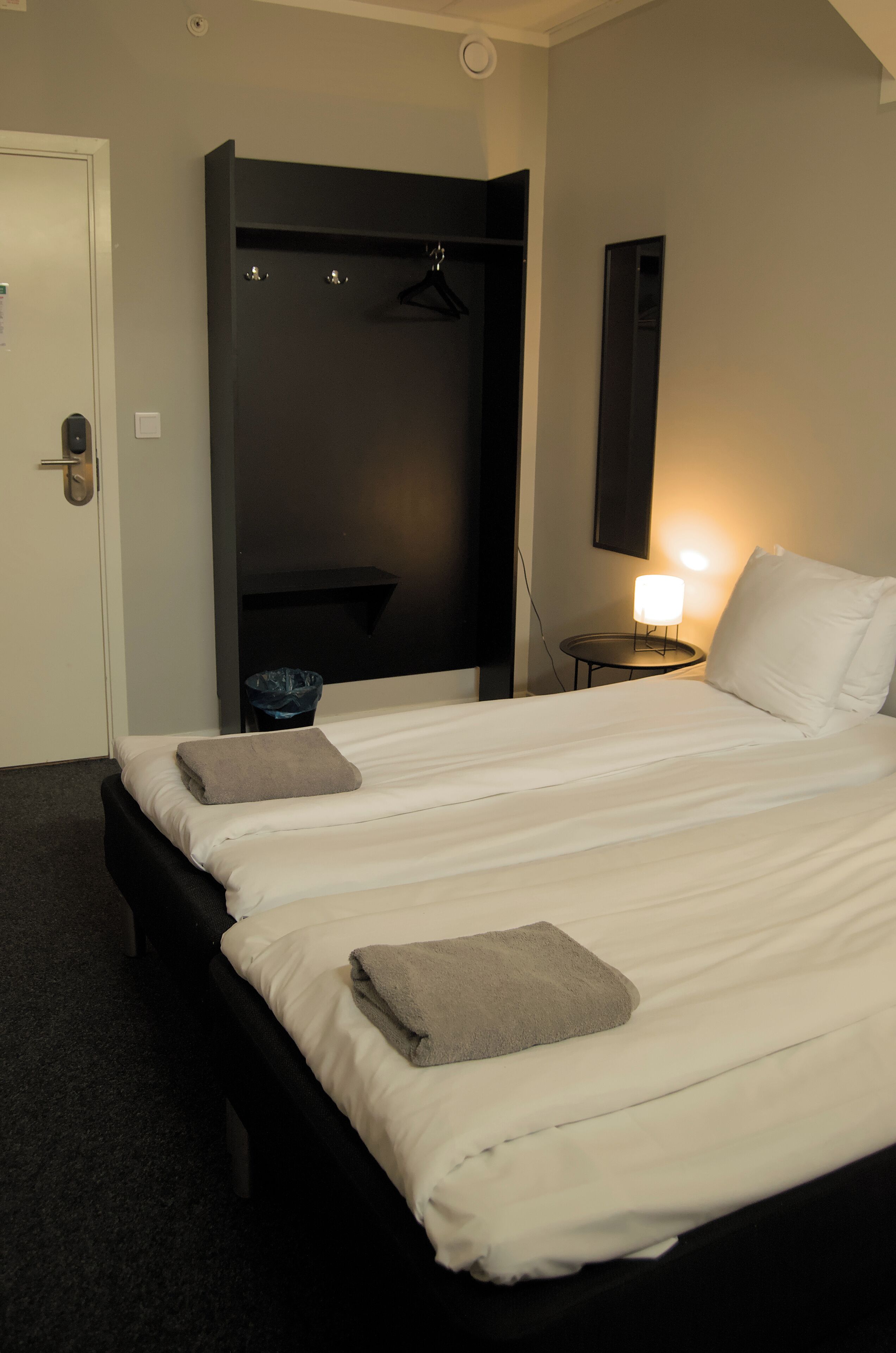 A hotel room with two beds, a wardrobe, a bedside table with a lamp, and a trash bin.