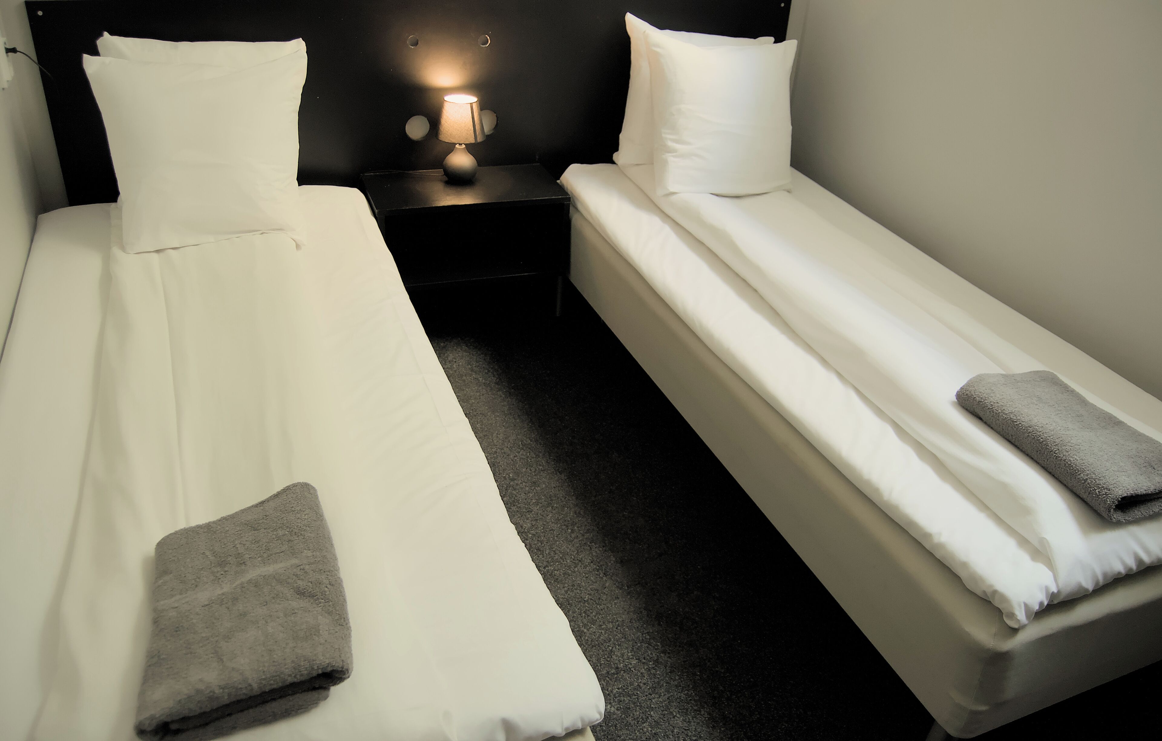 A hotel room with two single beds, a nightstand with a lamp, and a grey towel on each bed.