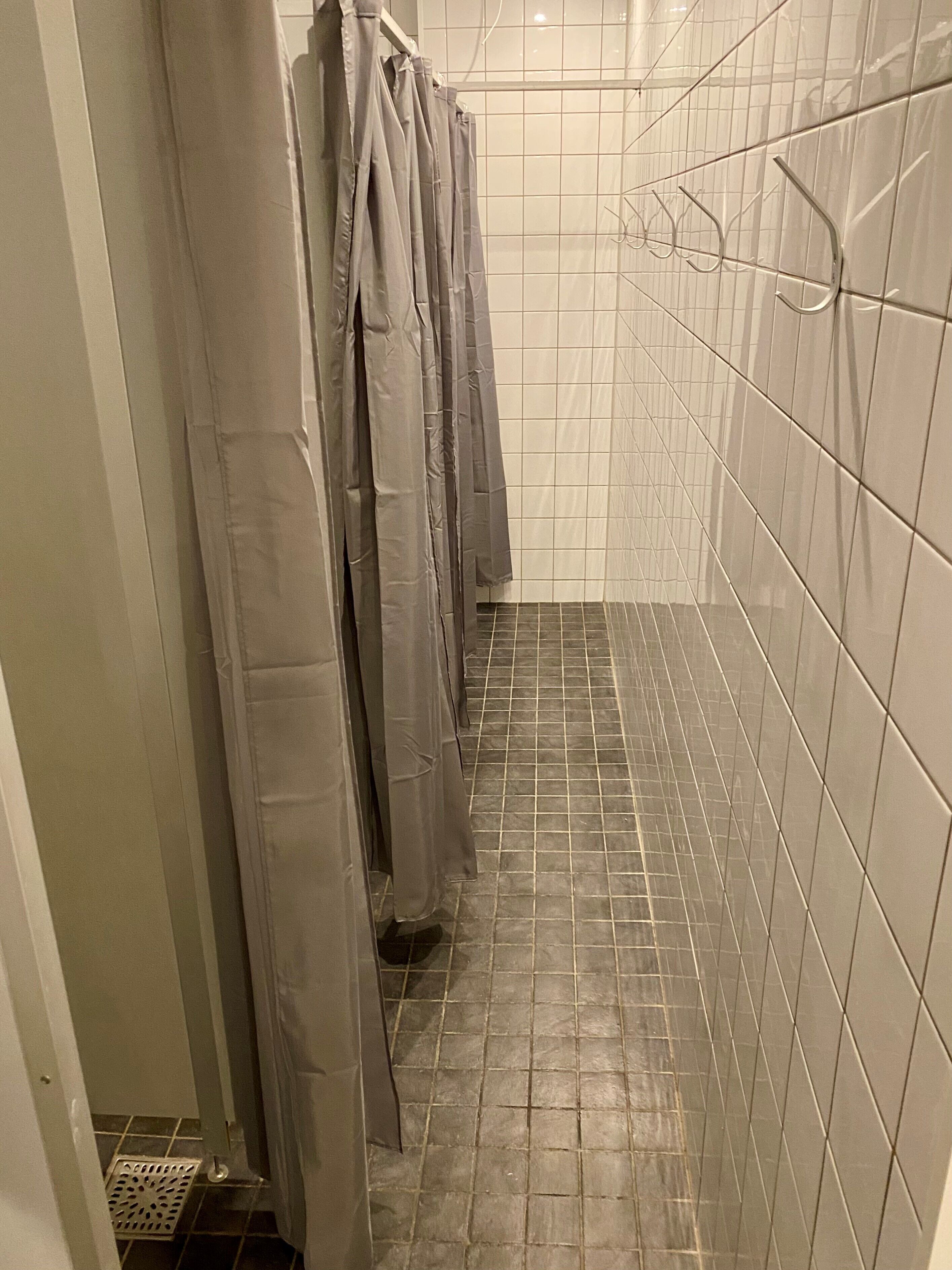 A narrow bathroom with tiled walls and floor, a metal shower drain, and a row of hanging shower curtains.