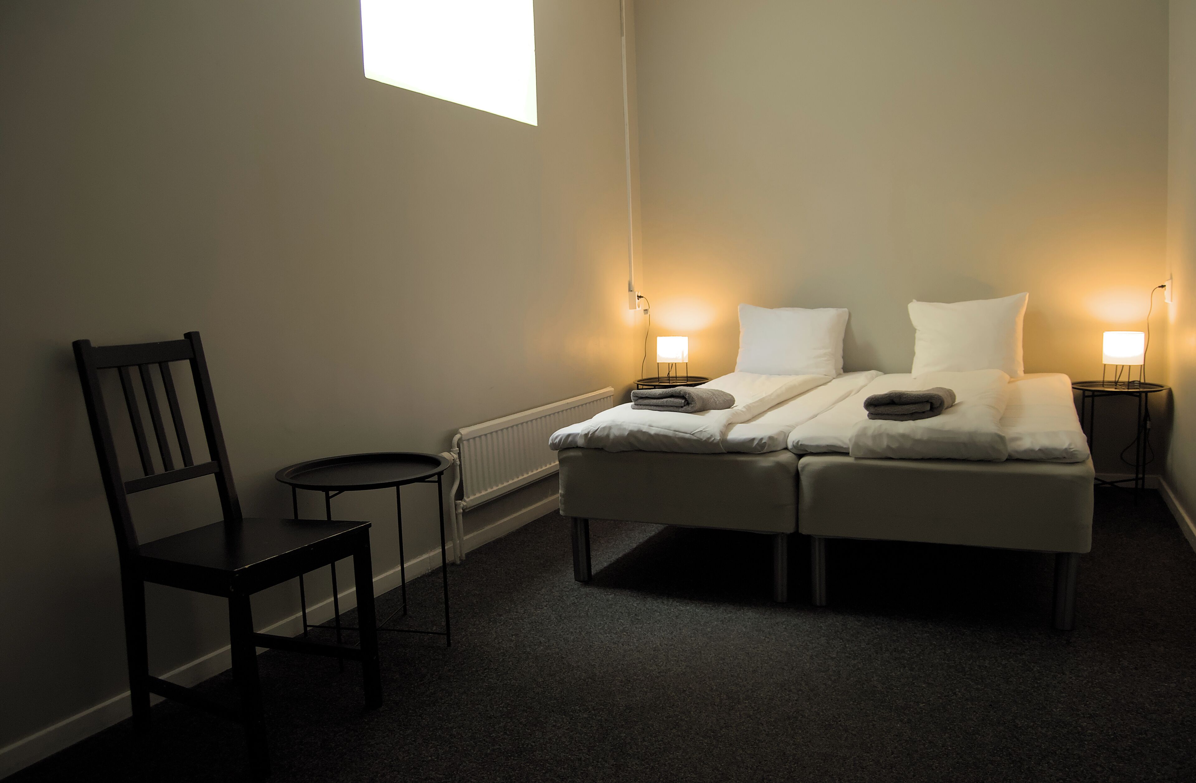 Um quarto pequeno e pouco iluminado, com uma cama de solteiro, duas mesas de cabeceira, uma cadeira e um radiador.
