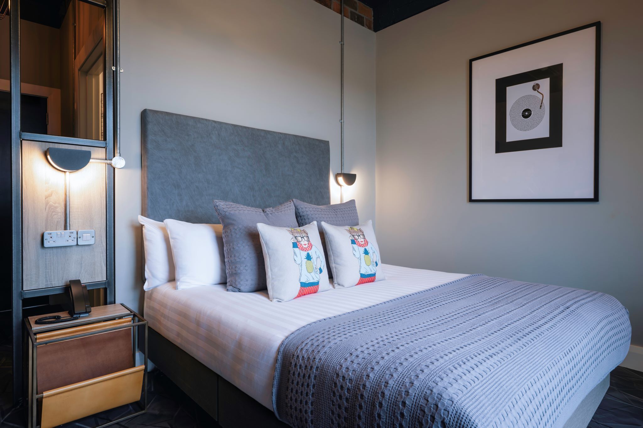 Cama bem arrumada com cabeceira cinza, almofadas brancas e manta tricotada cinza. Há uma moldura na parede e uma mesa de cabeceira com abajur.