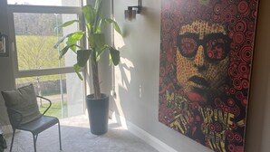 Ein Zimmer mit einem großen Fenster, einem Blumentopf, einem Sessel und einer Wand mit einem farbenfrohen abstrakten Gemälde.