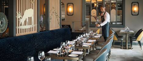 A restaurant with a long table set for dining, a waiter attending to guests, and a decorative wall with a crescent moon and antlers design.