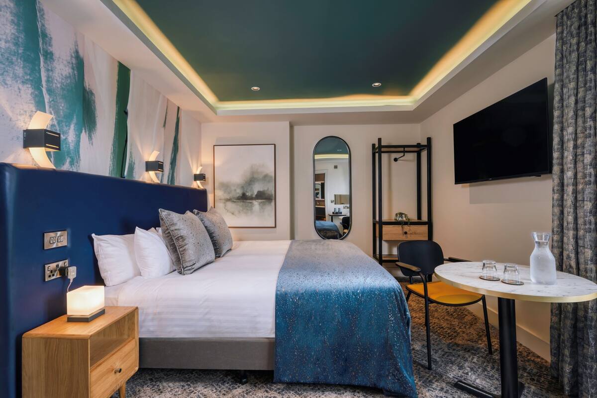 Una habitación de hotel moderna con una cama grande, lámparas de noche, un televisor de pantalla plana, una mesita con vasos y un espejo.