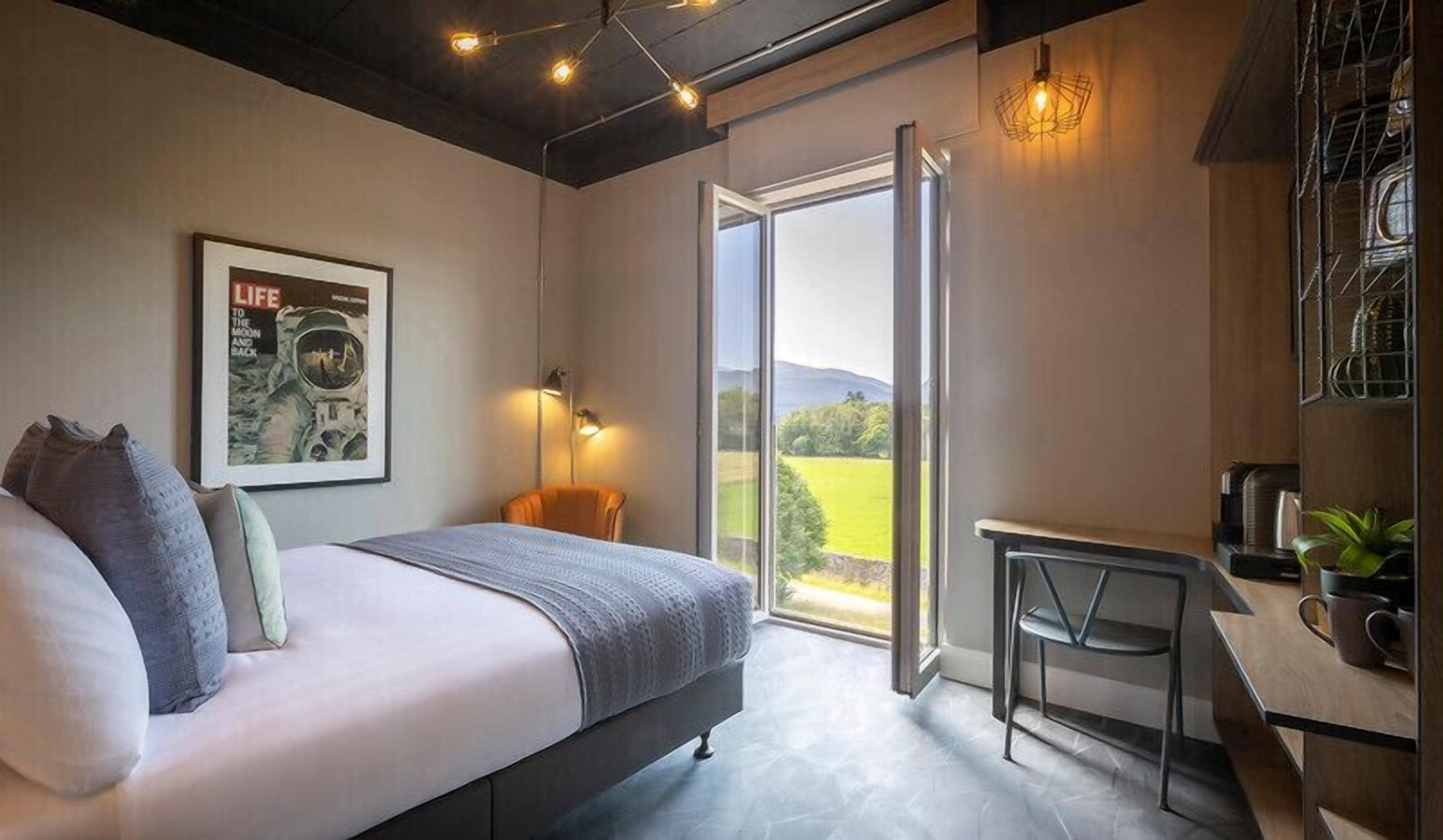 A bedroom with a bed, a desk, a chair, a window with a view of the countryside, and a wall-mounted light fixture.