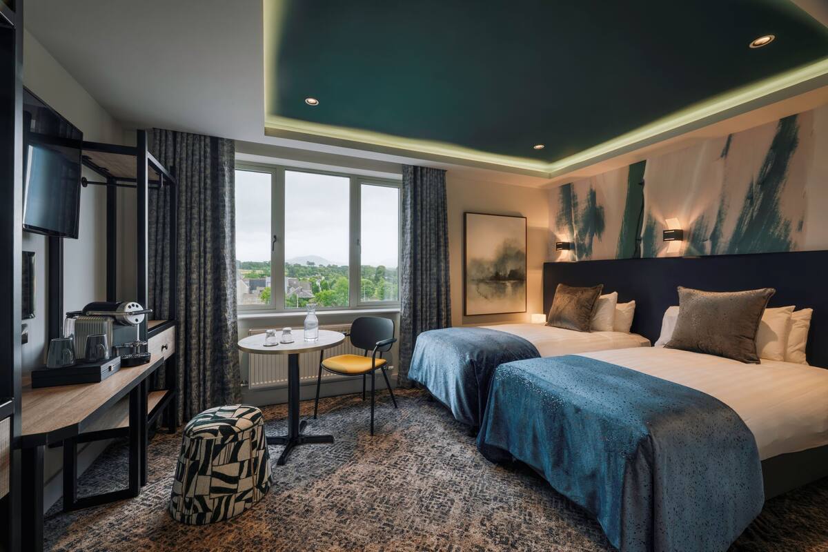 Habitación de hotel con dos camas, un escritorio con silla, una mesita y vistas a zonas verdes.