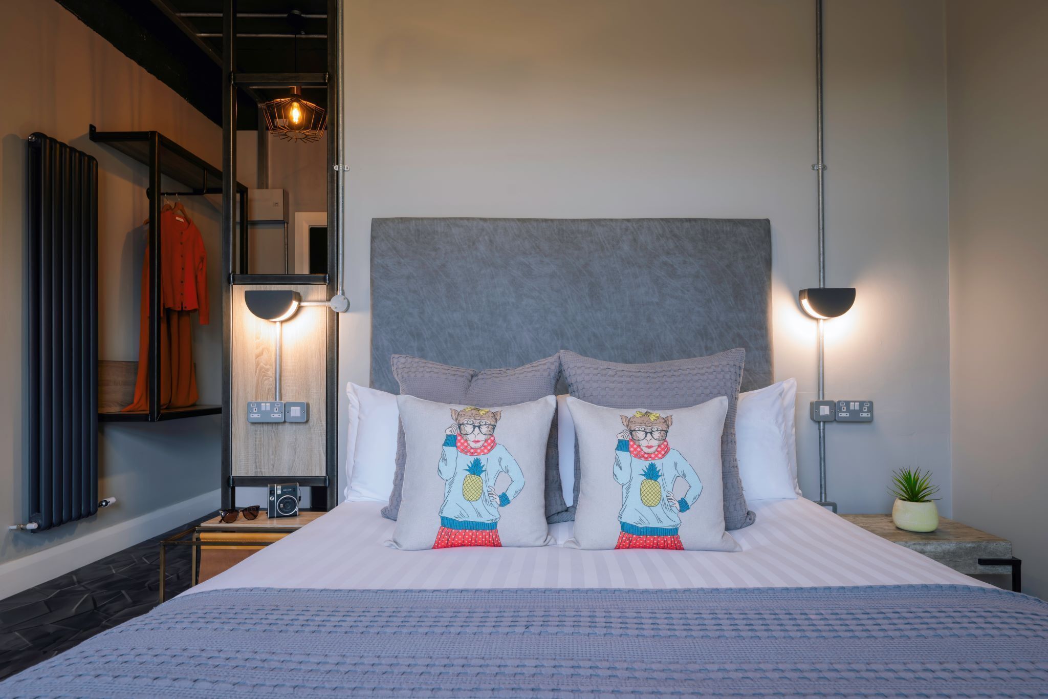 A neatly made bed with two decorative pillows, a bedside table with a radio, and a wall-mounted shelf with a jacket.