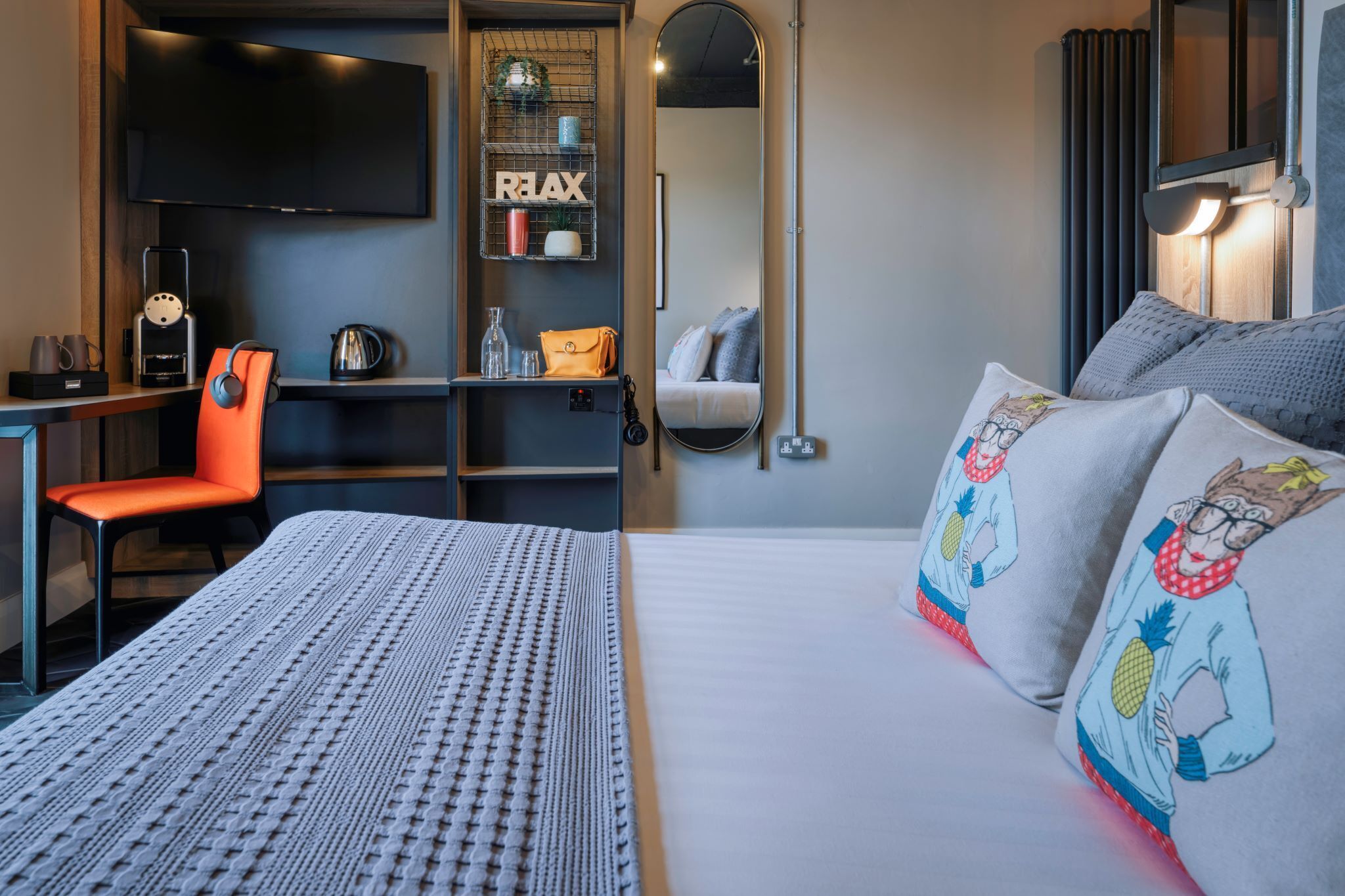 A hotel room with a bed, a desk with a chair, a TV mounted on the wall, and a shelf with various items.