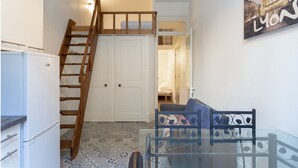 Een compacte kamer met een glazen tafel, een houten trap, een witte koelkast en een bed op de achtergrond.