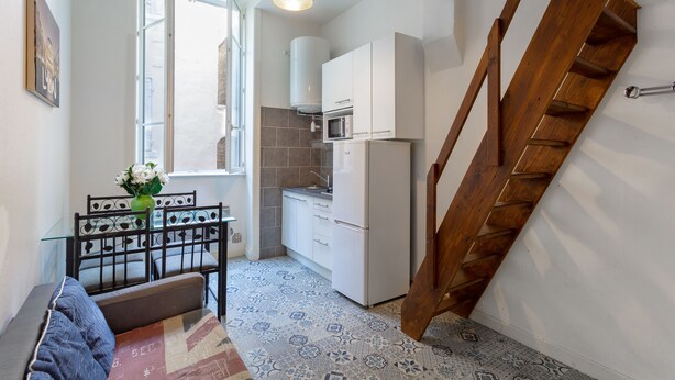 Een compacte keuken met een koelkast, magnetron en witte kasten. Er staat een eettafel met stoelen bij en een vaas met bloemen. Een houten trap is aan de rechterkant te zien.
