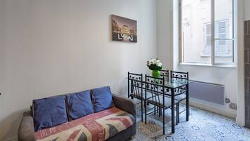 Een woonkamer met een bank, een eettafel met stoelen, een radiator en een raam met luiken.