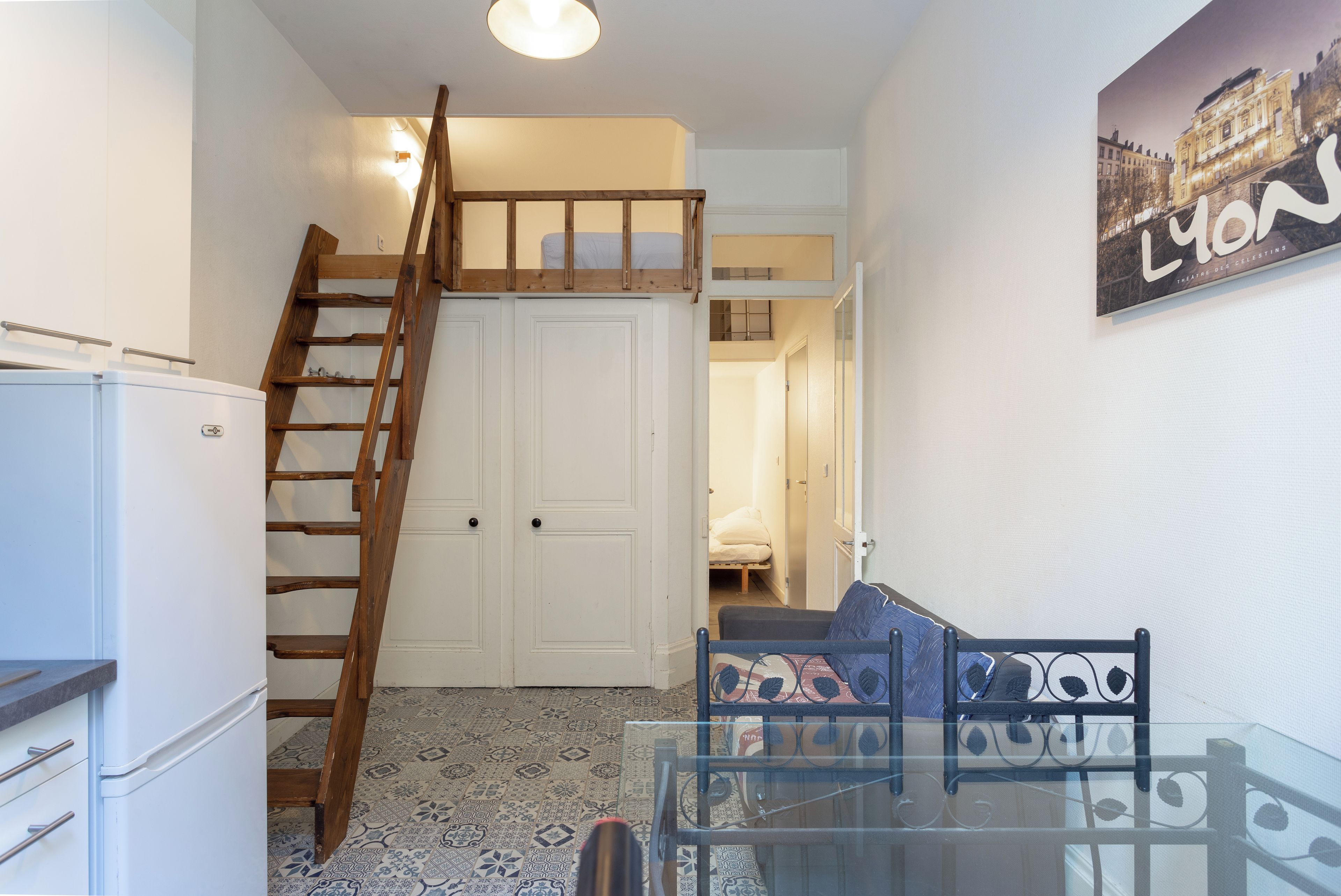 A compact room with a glass table, a wooden ladder, a white refrigerator, and a bed in the background.