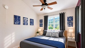 A bedroom with a bed, a ceiling fan, a window with curtains, and wall art.