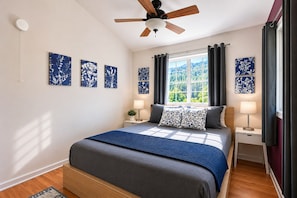 A bedroom with a bed, a ceiling fan, a window with curtains, and wall art.