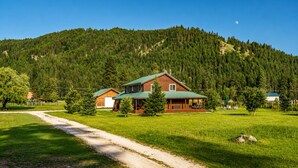 A cabin with a green roof, surrounded by a forested hillside, a dirt path, and a clear sky.