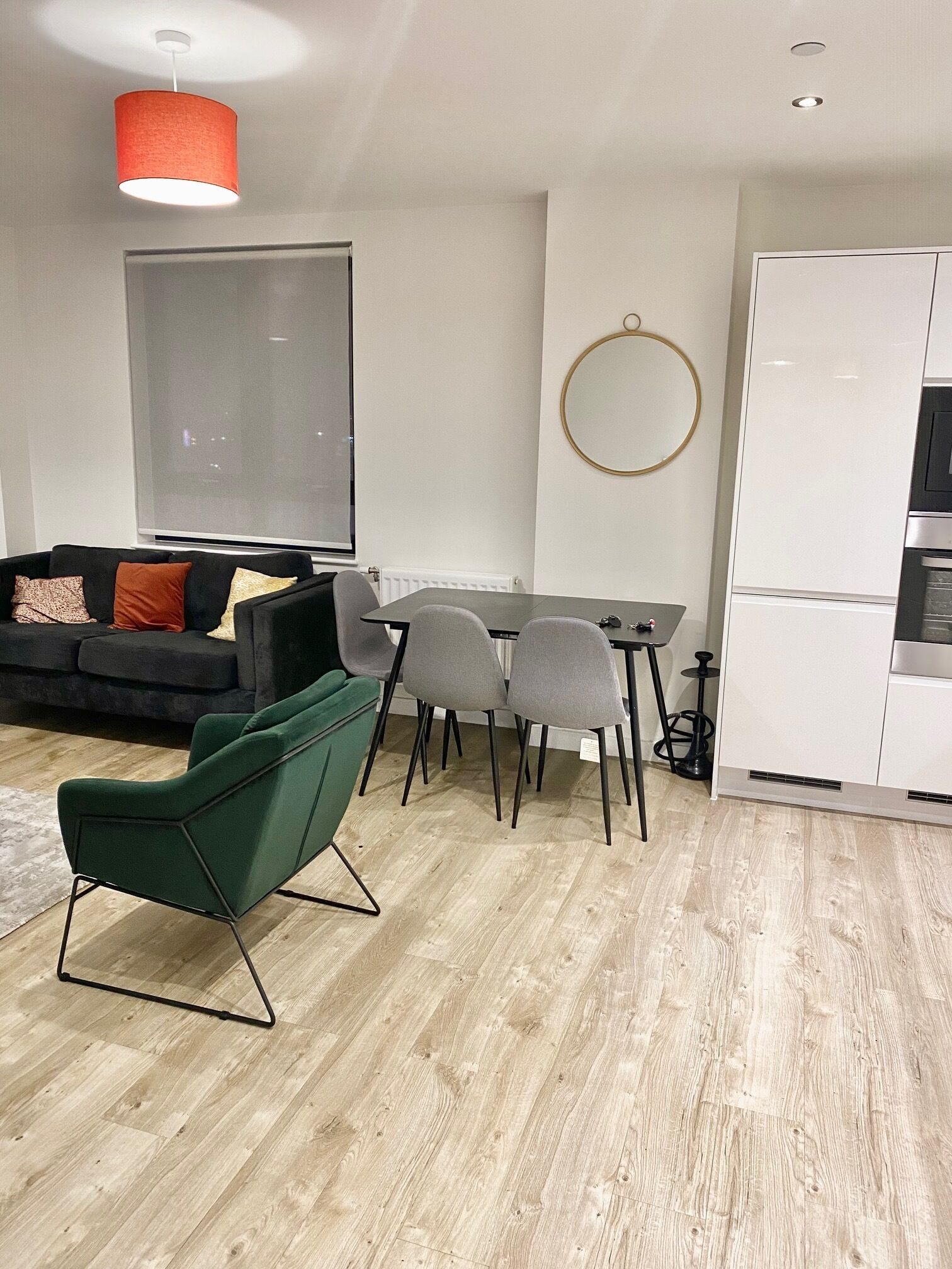 A modern living room with a sofa, dining table, and chairs. There is a large window, a round mirror, and a white cabinet.