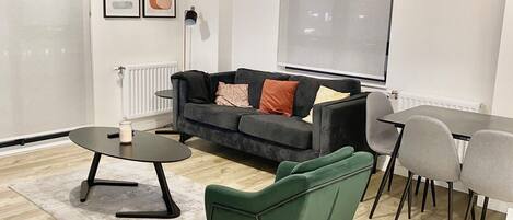 A modern living room with a black sofa, green armchair, and a dining table with chairs. There are two pendant lights, a radiator, and framed artwork on the wall.