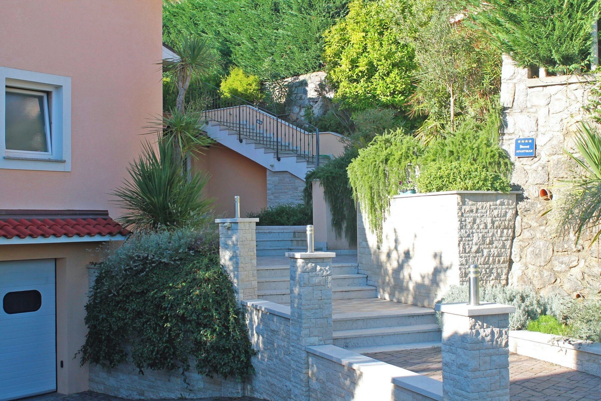 Un immeuble résidentiel doté d’un escalier en pierre menant à l’entrée du deuxième étage, entouré d’une végétation luxuriante et d’un mur en pierre.