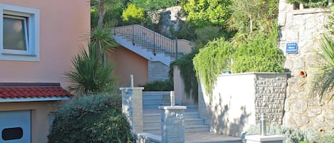 A residential building with a stone staircase leading to a second-floor entrance, surrounded by lush greenery and a stone wall.