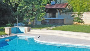 A swimming pool with a tiled edge, a stone pathway, and a house with a red-tiled roof in the background. - Bruk in Lovran - Kvarner Region / Bruk in Lovran - Appartement für 2 Personen AP2 (Lovran)