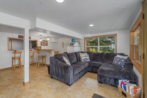 Ein modernes Wohnzimmer mit einem grauen Ecksofa, Holzstühlen, einer kleinen Küchenzeile und einem großen Fenster mit Blick ins Grüne.