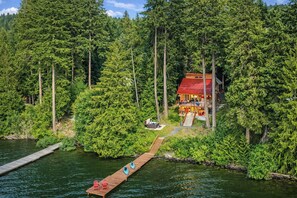 Une cabane entourée d’une forêt dense, un quai menant à la cabane et un petit bateau sur l’eau.