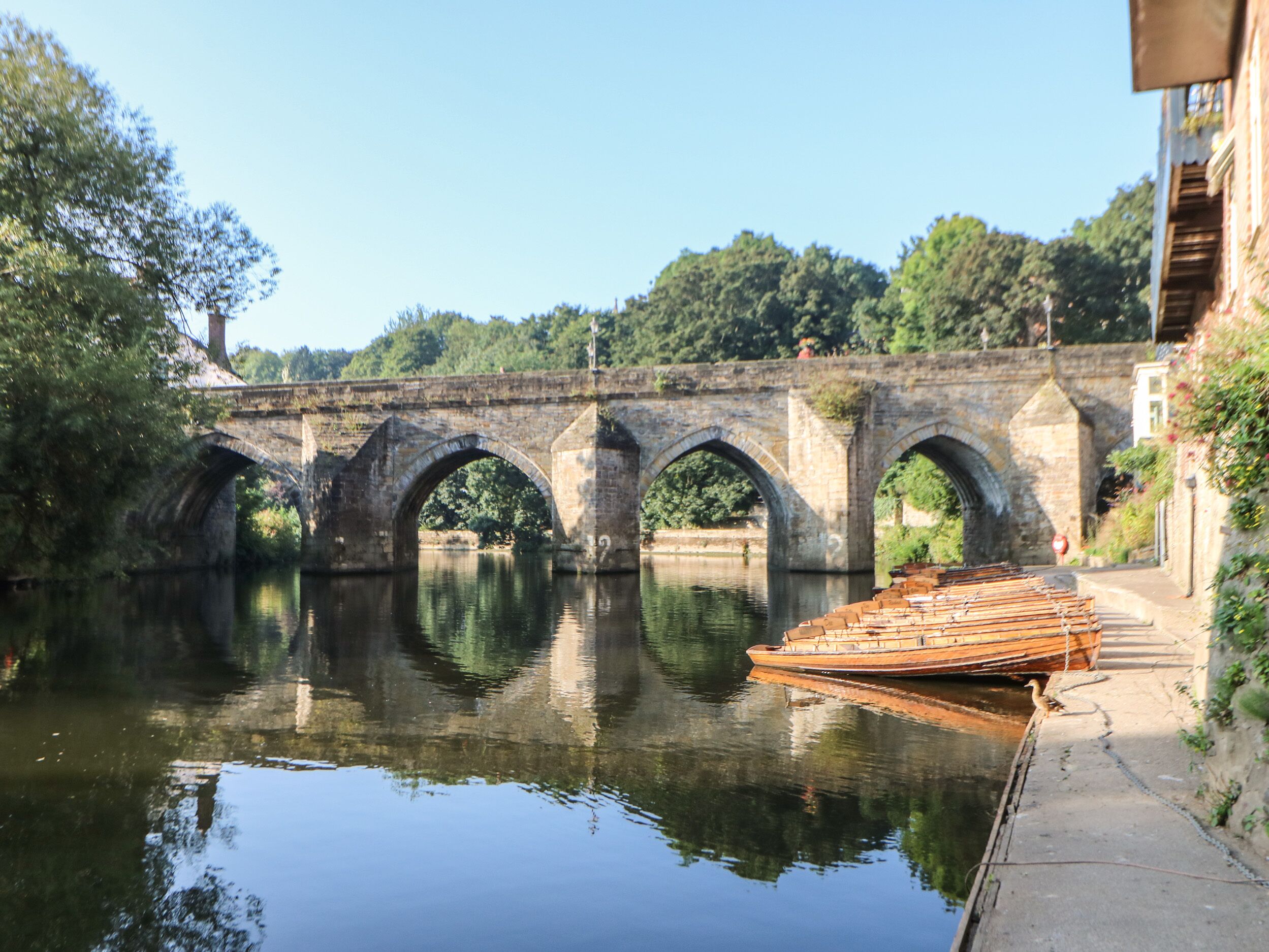 Eine Steinkonstruktion überspannt einen ruhigen Fluss, rechts ist ein kleines Boot vertäut.