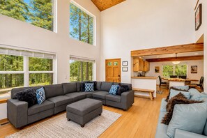 A spacious living area with a large sectional sofa, a small ottoman, and a light blue couch. There is a wooden dining table with chairs, a kitchen area with wooden cabinets, and large windows offering views of trees. - Just Plain Getaway 3 Bedroom Home by NW Comfy Cabins by RedAwning (Leavenworth)