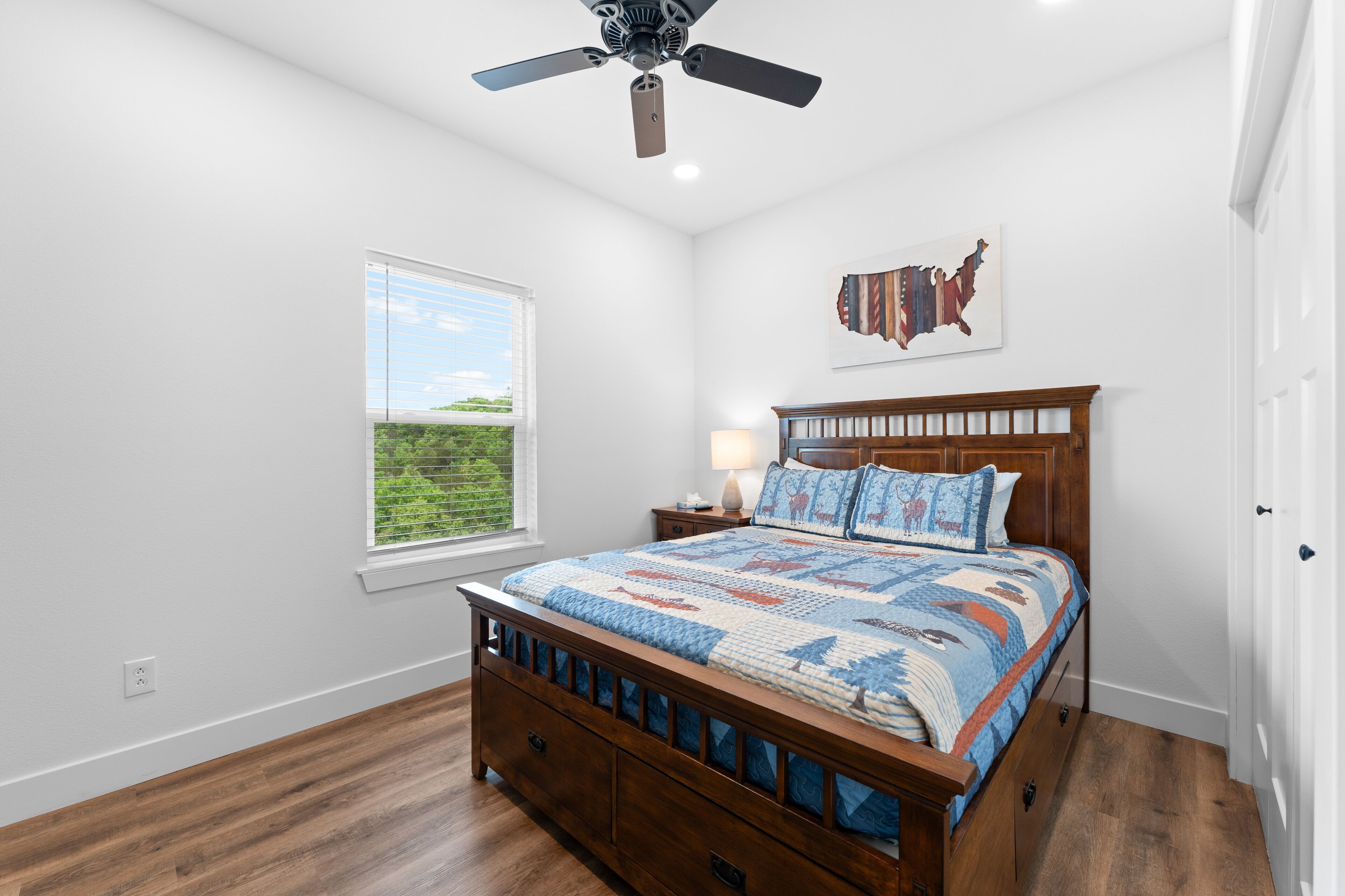 A bedroom with a wooden bed, a ceiling fan, a window with a view of greenery, and a wall map.