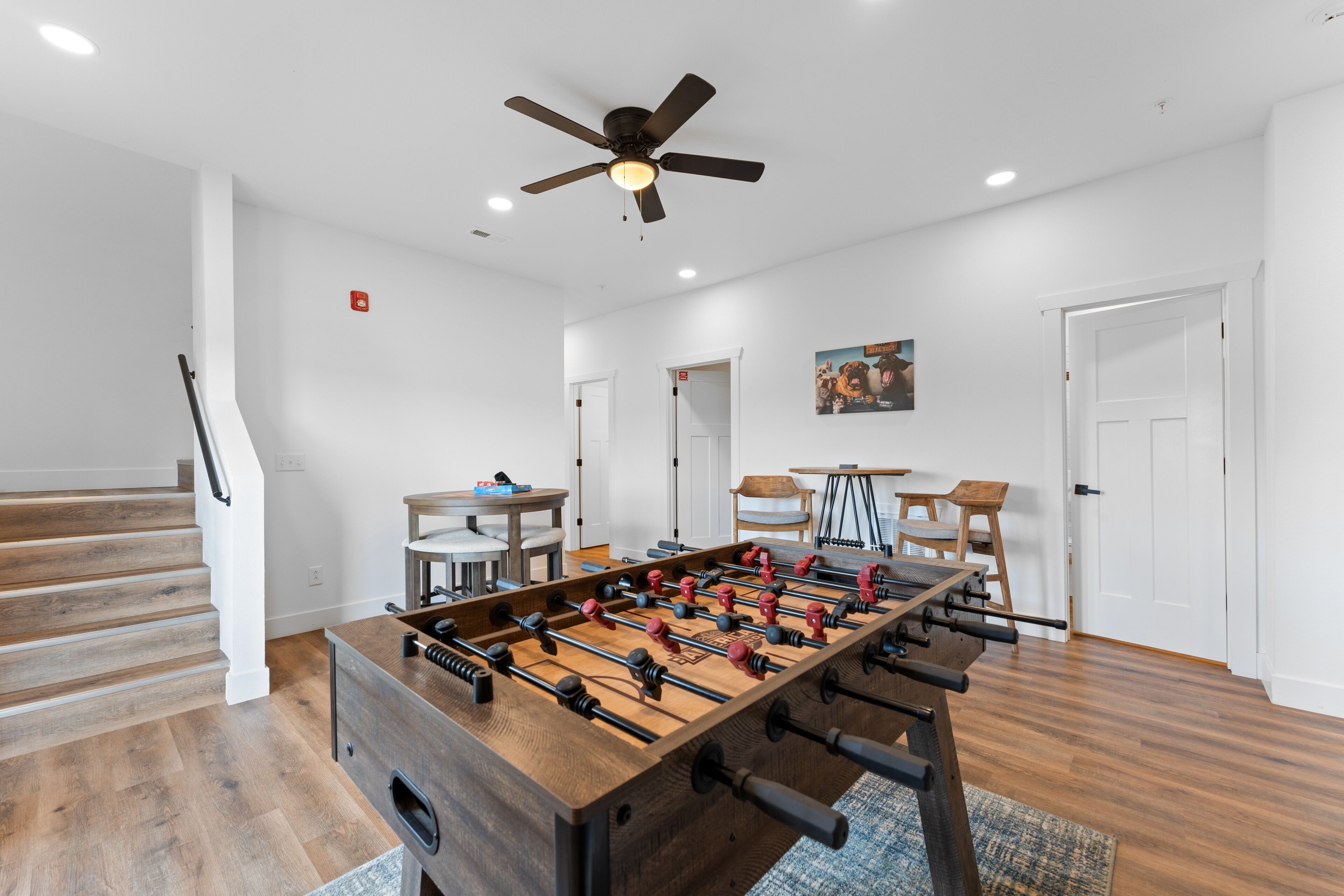 A spacious room with a foosball table, wooden flooring, a ceiling fan, and a staircase.