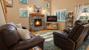 Una sala de estar acogedora con chimenea de piedra, sofás de cuero, un televisor y una estantería.