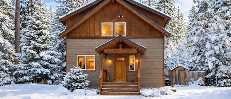 A wooden cabin with a front porch, surrounded by snow-covered trees.