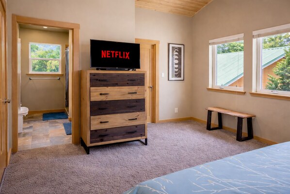 A bedroom with a wooden dresser, a bench, a bed, a door leading to a bathroom, and a window with a view of trees.