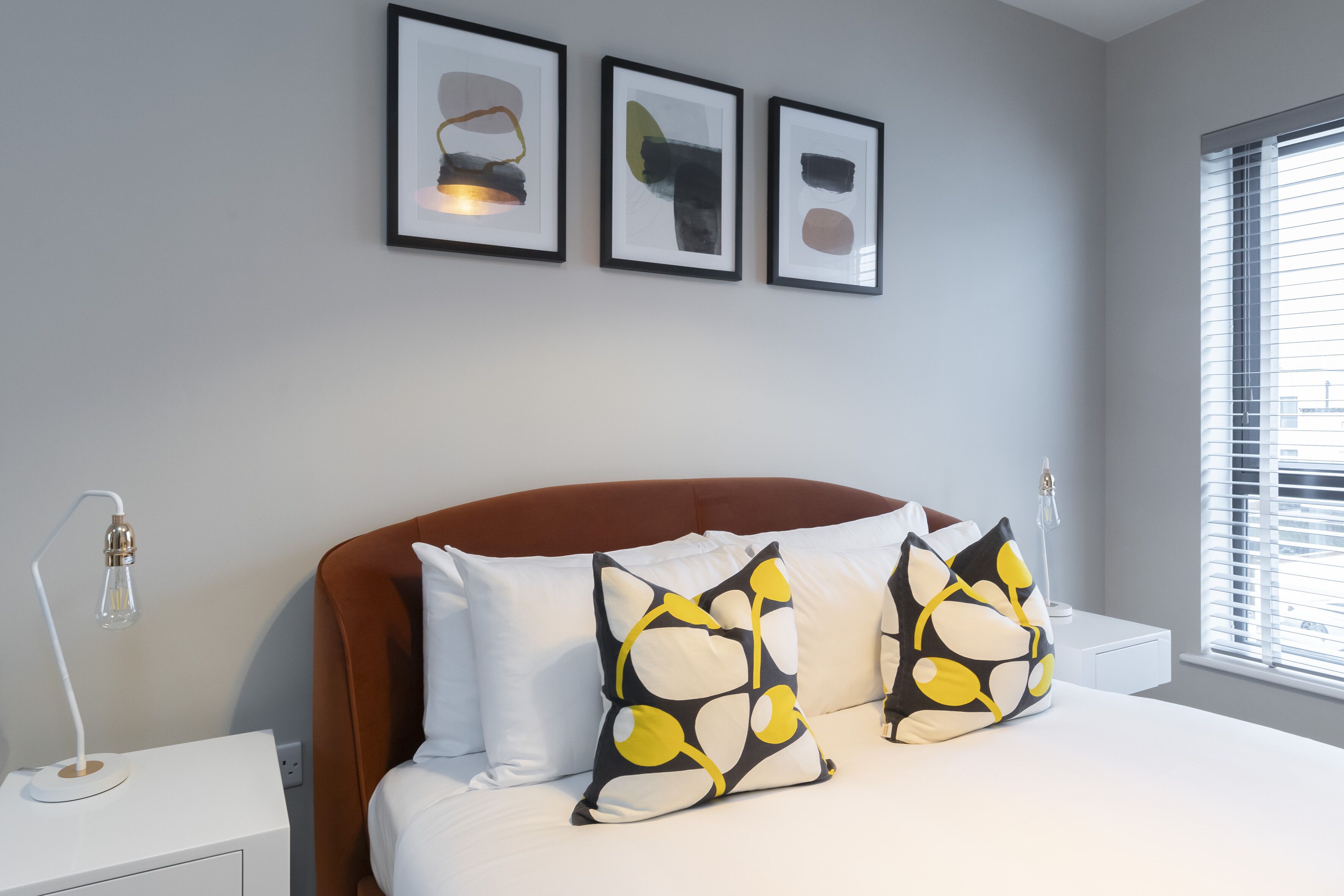 A bedroom with a bed, two bedside lamps, a nightstand, and three framed artworks on the wall.