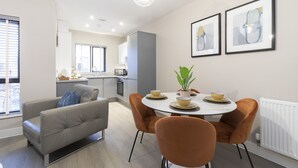 Een moderne woonkamer met een grijze bank, oranje fauteuils, een ronde eettafel met kommen en een plant, en een keuken op de achtergrond.