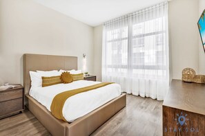A hotel room with a bed, a nightstand, a wooden desk, and a large window with sheer curtains.