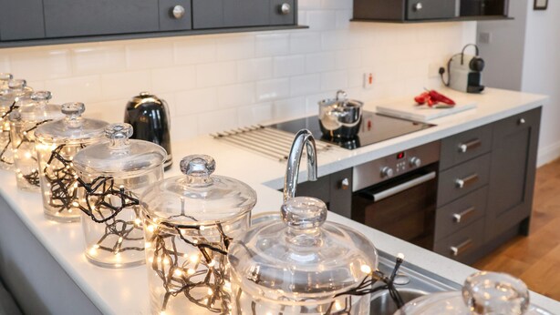 A modern kitchen with grey cabinets, a white countertop, and glass jars with string lights. - Ebenezer Chapel (Saxmundham)