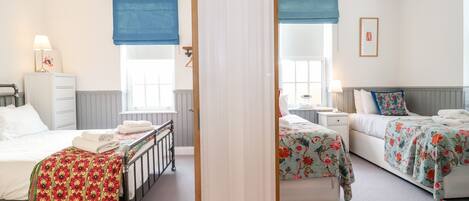A hotel room with two beds, a window with a blue blind, a bedside table with a lamp, and a wall-mounted clock.