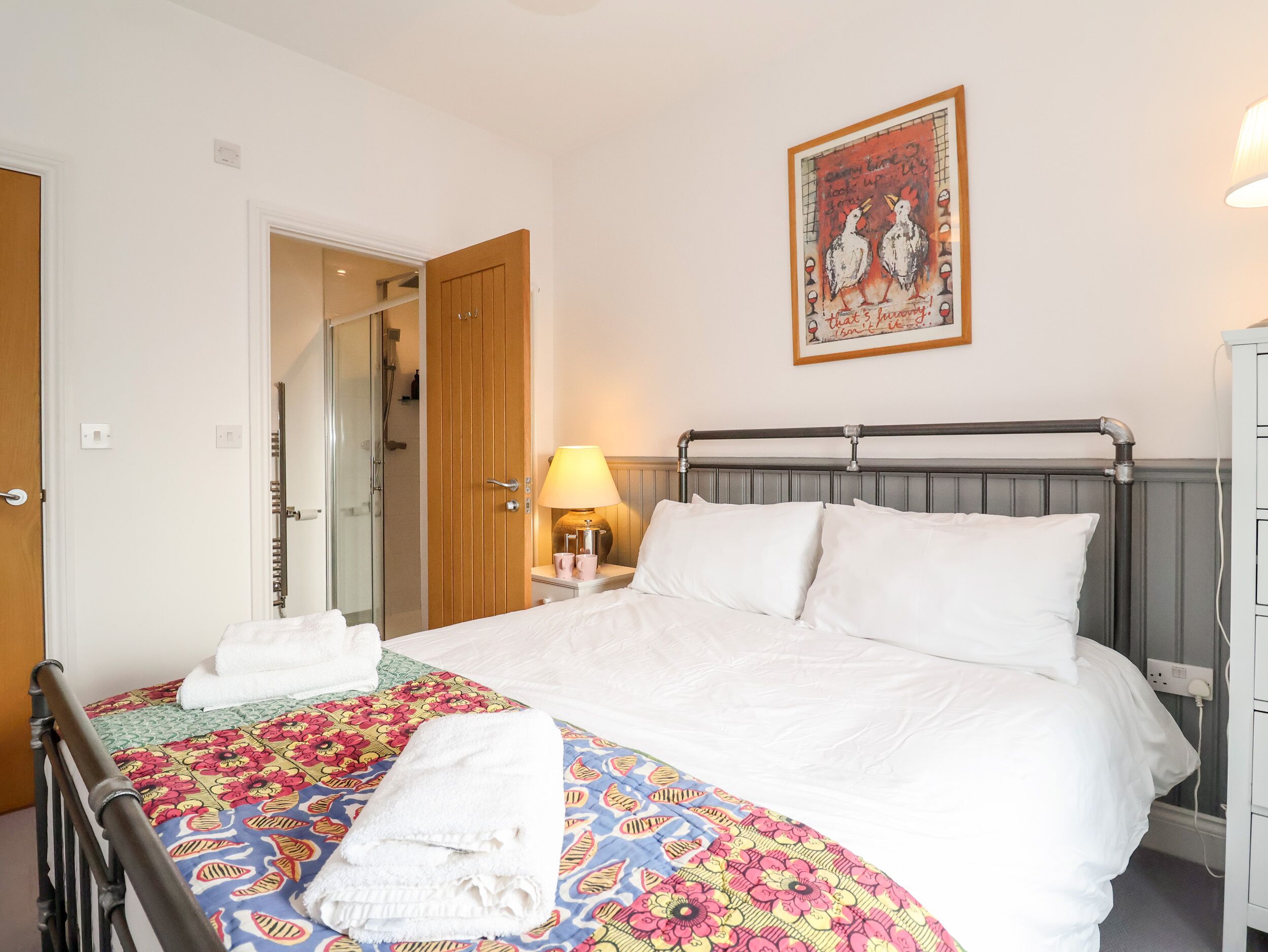 A neatly made bed with a floral-patterned bedspread, a wooden door, a bathroom with a glass shower door, and a wall-mounted picture.