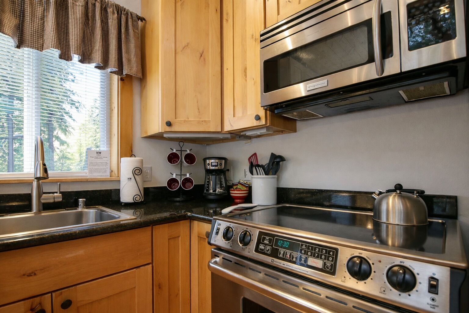 A kitchen with wooden cabinets, a stainless steel stove, a microwave, and a window with blinds.