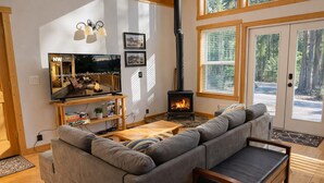 A cozy living room with a large gray sofa, a wooden coffee table, a TV on a stand, a fireplace, and a large window with blinds. - Cascades Adventure Cabin 2 Bedroom Home by NW Comfy Cabins by RedAwning (Leavenworth)