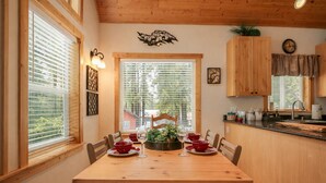 A dining area with a wooden table set for four, surrounded by chairs. There are two windows with blinds, a wooden ceiling, and a kitchen area with wooden cabinets and a sink. - Cascades Adventure Cabin 2 Bedroom Home by NW Comfy Cabins by RedAwning (Leavenworth)