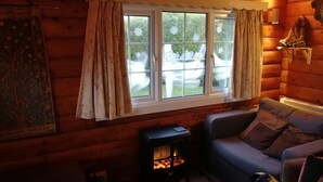 L’intérieur d’un chalet chaleureux, avec un mur en rondins de bois, un canapé, une petite table avec un foyer et une fenêtre agrémentée de rideaux.