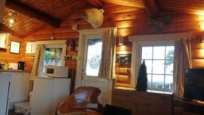 L’intérieur d’un chalet accueillant, avec un plafond en bois, une table à manger sur laquelle se trouve un ordinateur portable, une chaise en bois, un coin cuisine équipé d’un four à micro-ondes et une fenêtre avec des rideaux.