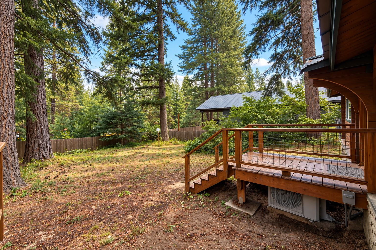 Une terrasse en bois avec un escalier menant à une zone abritée, entourée d’une forêt dense et d’une cour clôturée.