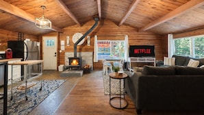 En mysig stuga med träpanel i taket, en öppen spis, ett köksutrymme, ett vardagsrum med en soffa och en tv som visar "NETFLIX".