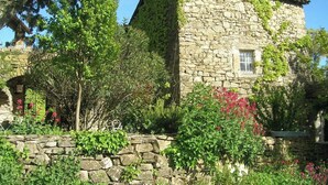 A stone building with a tiled roof, surrounded by lush greenery and flowering plants. - Charming detached house with terrace in a farmhouse with swimming pool and garden (Vesseaux)